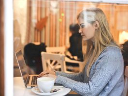 7 Ways To Organize Your OnlyFans Feed Like A Pro Blonde woman in a blue sweater using a laptop in a cozy café setting.