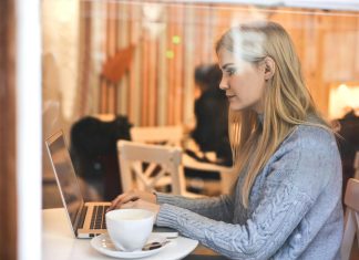 7 Ways To Organize Your OnlyFans Feed Like A Pro Blonde woman in a blue sweater using a laptop in a cozy café setting.