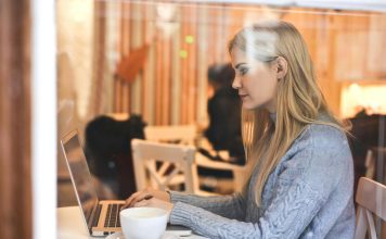 7 Ways To Organize Your OnlyFans Feed Like A Pro Blonde woman in a blue sweater using a laptop in a cozy café setting.