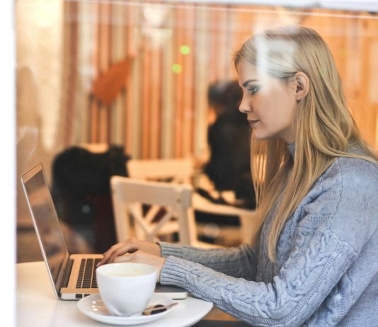 7 Ways To Organize Your OnlyFans Feed Like A Pro Blonde woman in a blue sweater using a laptop in a cozy café setting.